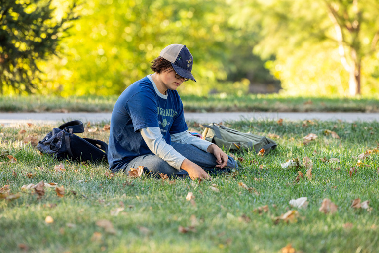 student sitting outside in the grass with blue shirt and hat