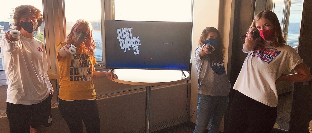 Four students holding game controllers with a TV that has text that reads "Just Dance 3" in the background