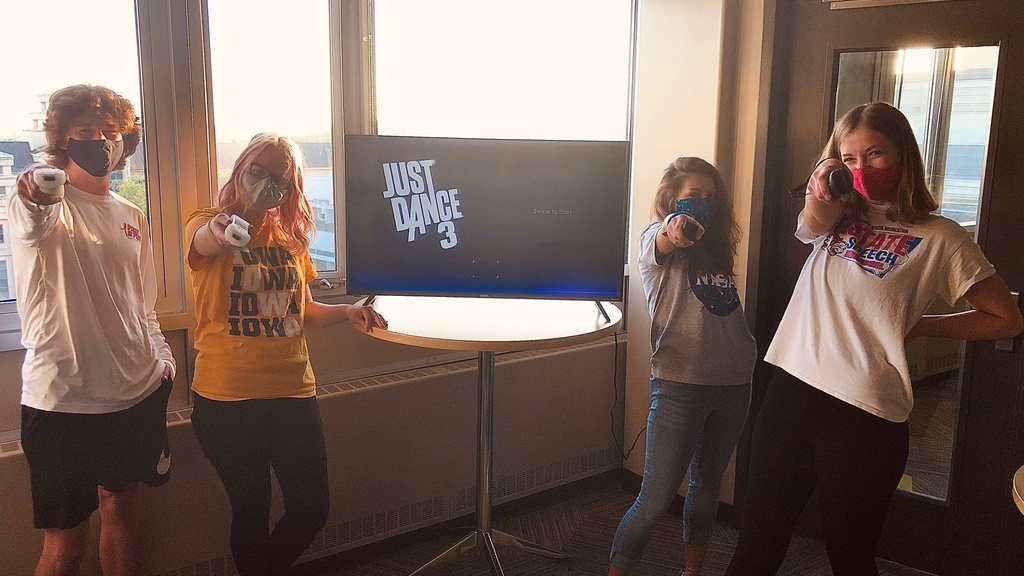 Four students holding game controllers with a TV that has text that reads "Just Dance 3" in the background